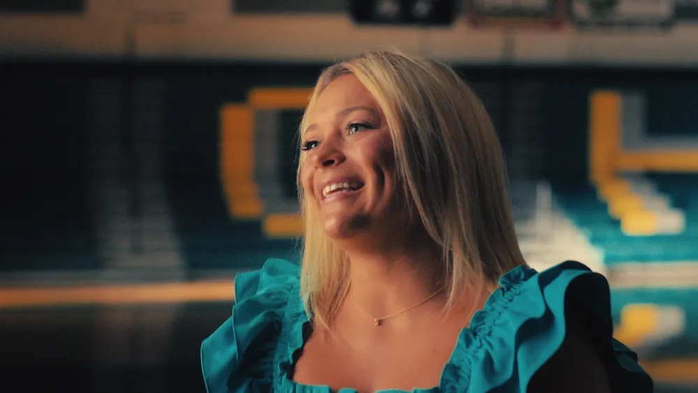 A smiling blonde woman in a teal top in front of a gym wall with a large yellow letter