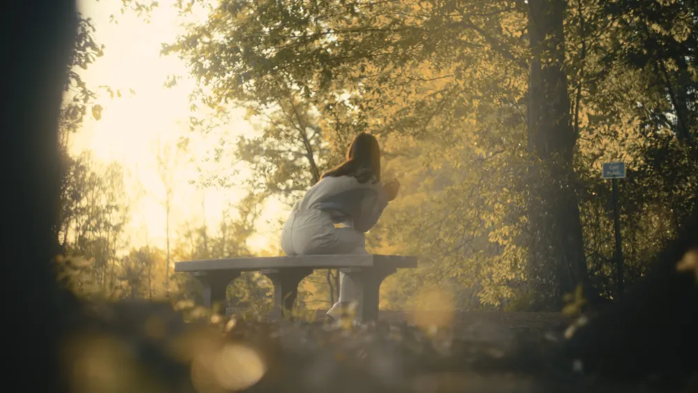 A woman seated on a stone bench in a sunlit forest beside a pond